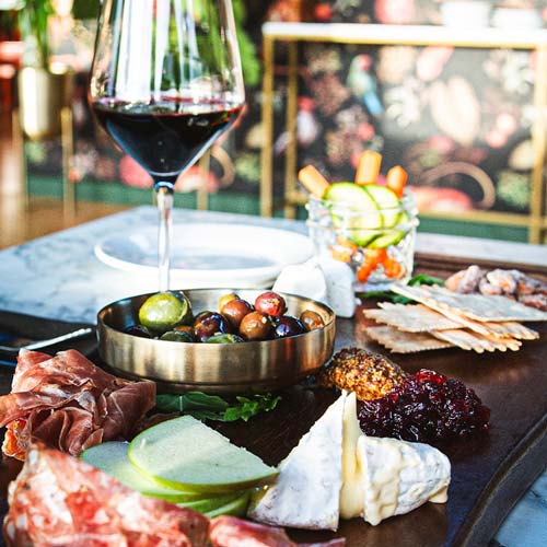 A charcuterie board with olives, green apple slices, brie, crackers, prosciutto, and a glass of red wine